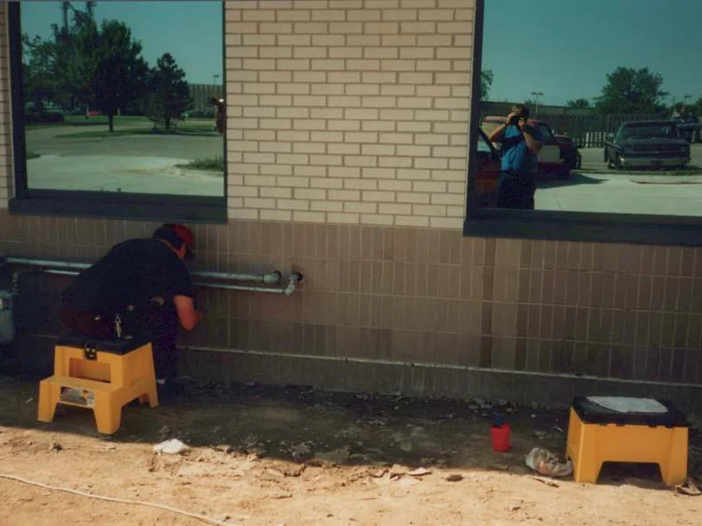 An applicator crouched on a yellow step stool working stain into the tan base course beside a conduit in 1995, the photographer reflected in the window above