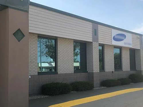The long elevation of the building from the parking lot in 2019, cream brick over a dark base under a band of siding, a tenant sign at the right and a yellow curb line in front