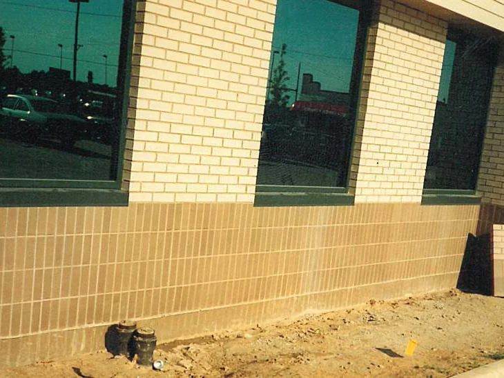 Three window bays in 1995, cream brick above a tan base course, bare dirt at the foot of the wall and a stack of sample brick leaning against it