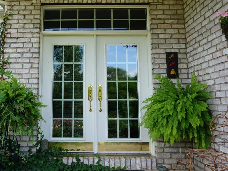 The front entry in 2019, white double doors between two ferns, the brick either side one even buff range with a wall planter to the right