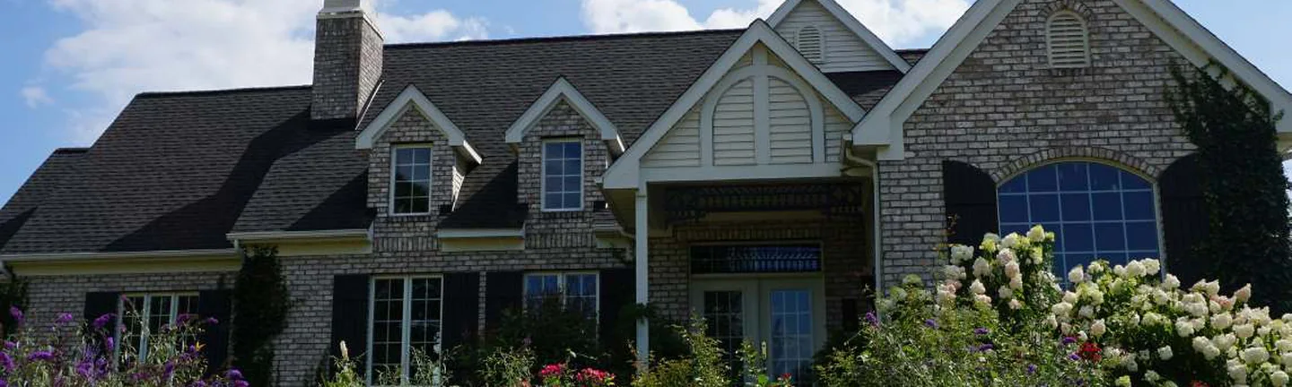 The front of the house in 2019, buff brick under a dark shingle roof above a full bed of hydrangeas and perennials