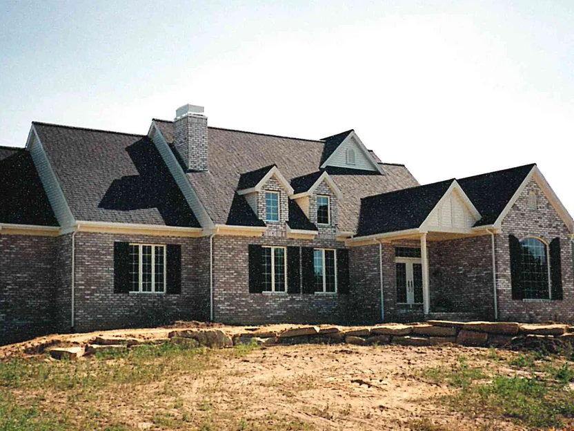 The front of the house in July 1997, newly built on bare dirt, its grey and tan brick mottled with dark units under a dark shingle roof with three dormers