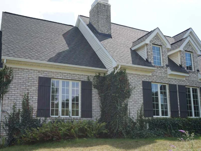 The side elevation in 2019, two dormers above shuttered windows, ivy climbing the buff brick between them and ferns along the base