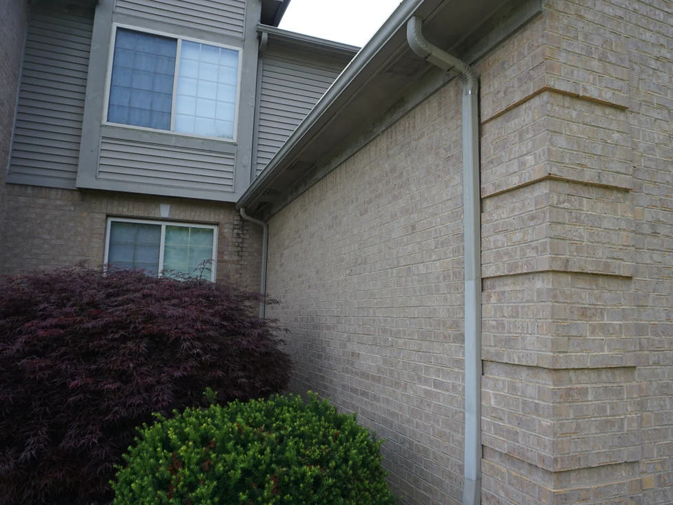An outside corner of the house in 2019 with a downspout running down it, the brick holding one buff and tan range across both faces below the siding
