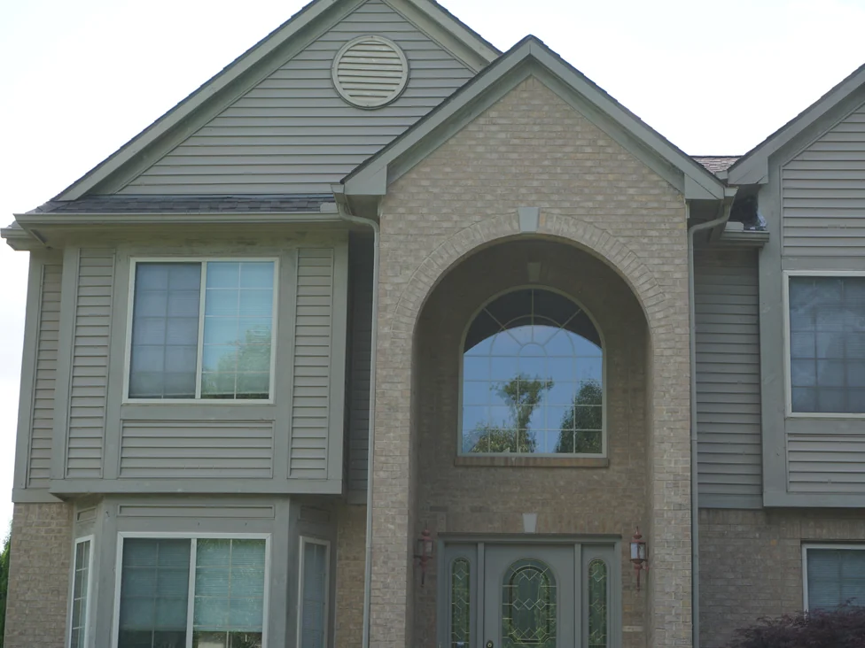 The front of the house close up in 2019, the arched brick entry running two storeys between sided gables with a leaded glass door beneath it