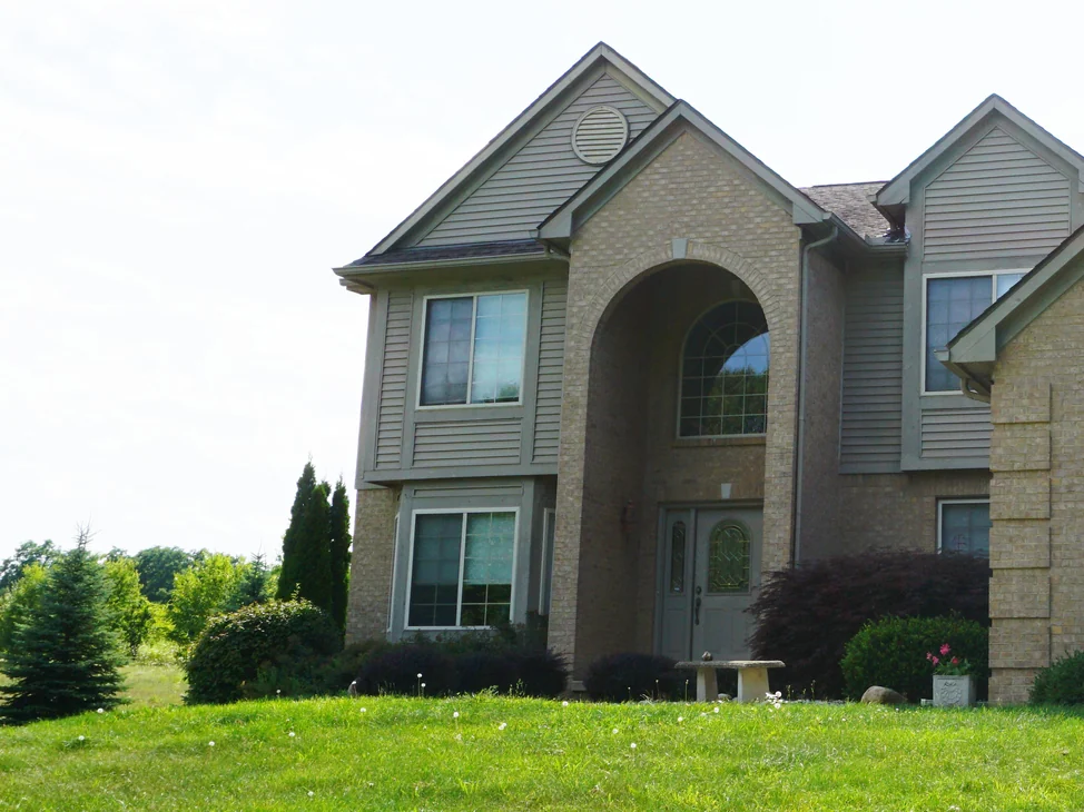 The house from its front lawn in 2019, the tall arched brick entry and the gabled wing beside it reading as one buff and tan range above clipped planting.