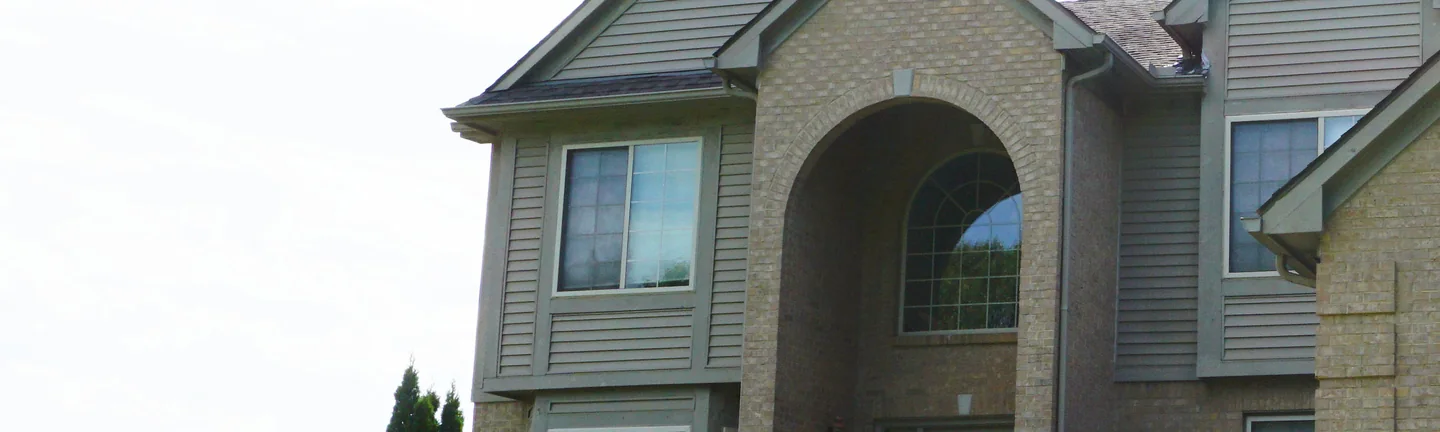 The front of a two-story brick house in 2019, its arched entry and gable reading as one buff and tan range above mature planting