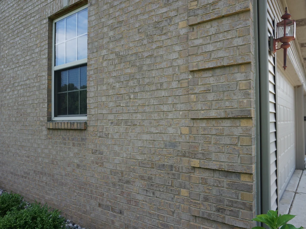A side wall of the house in 2019 seen at a low angle, projecting courses of tan and gold brick running across a grey and buff field toward the garage