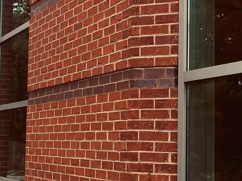 The same 135 degree corner in 2019, the corner shapes and the stretcher brick reading as one red range between two dark accent courses, windows either side reflecting trees and ivy climbing the base