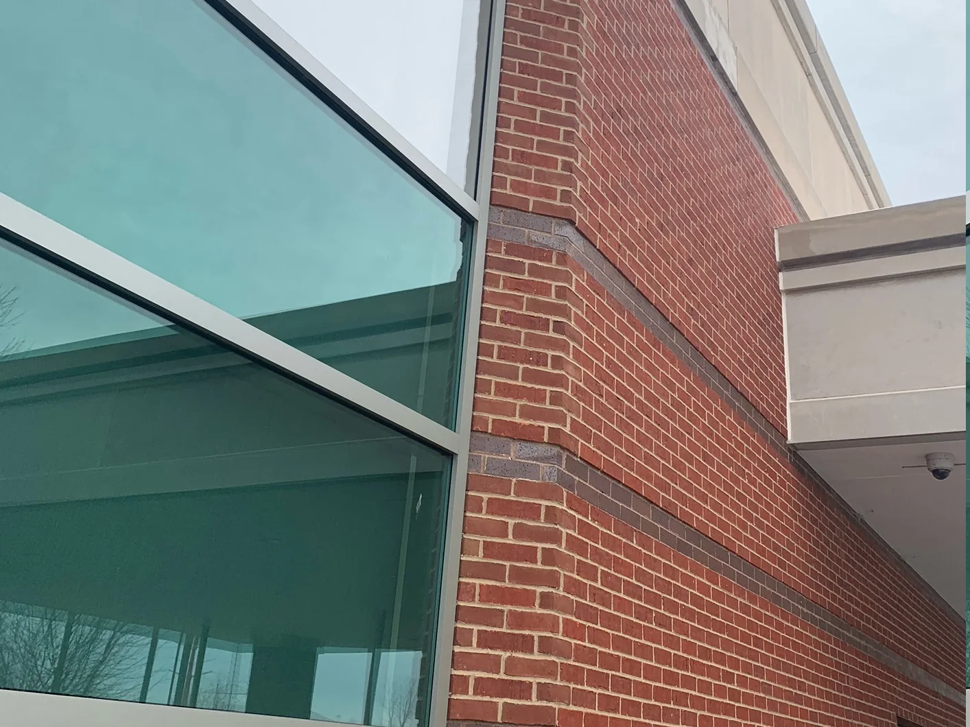 Looking up the corner in winter 2021, a glass curtain wall on the left and the red brick corner with its dark accent courses on the right, running up to a stone parapet under a grey sky