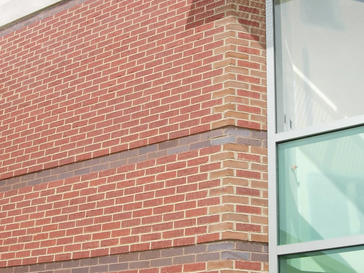 The 135 degree corner of the bank in 2004 before staining, seen from below in sunlight: red brick wall with two dark accent courses, the corner units a lighter pink than the stretchers, a stone band above and a glass window bay to the right