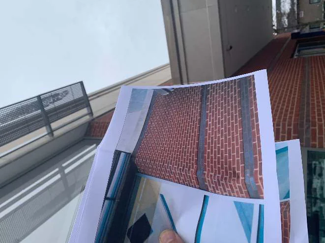 A hand holding a printed 2004 photograph of the corner up against the same corner in 2021, the soffit grille and sky behind