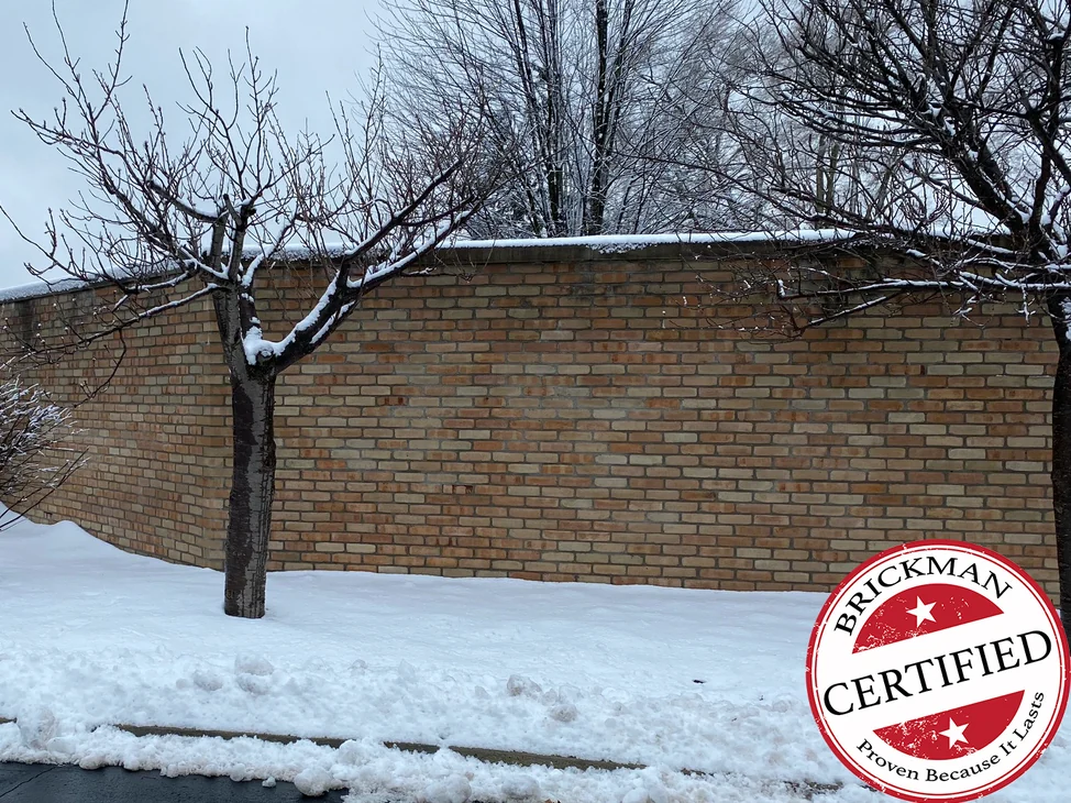 A long buff brick wall at the church in 2020, snow on its coping, a bare tree in front of it and the certification stamp in the corner of the frame