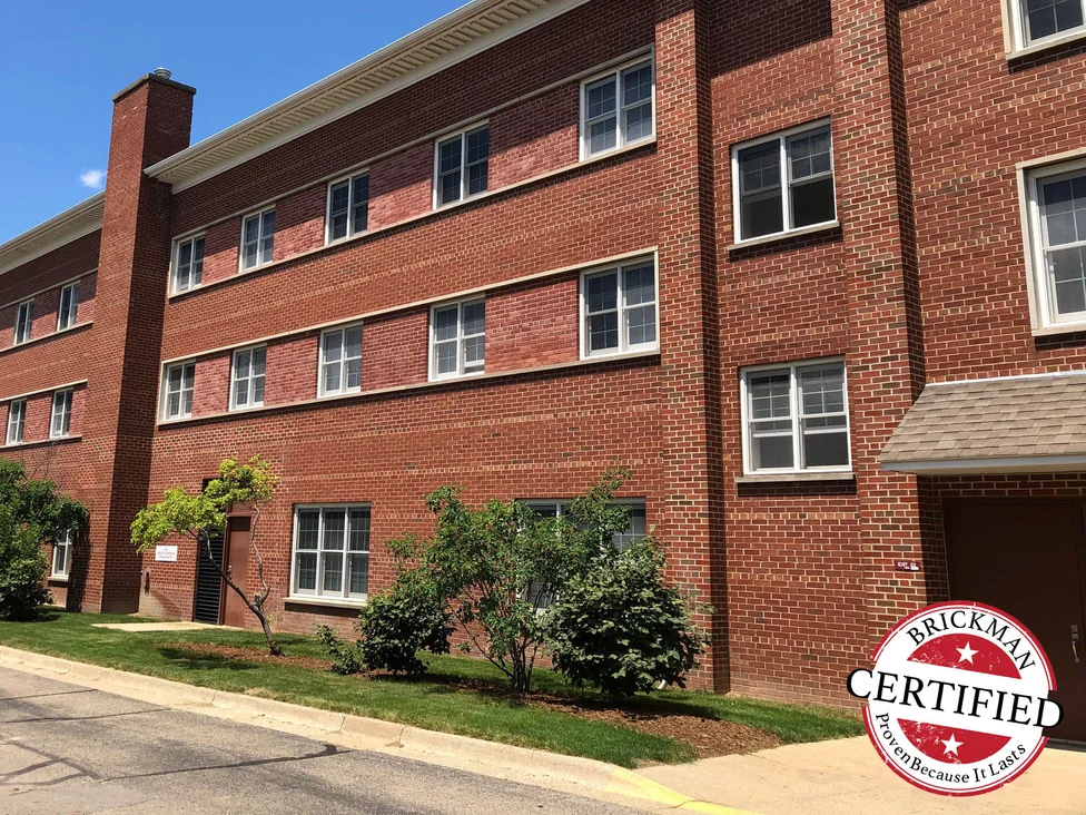 A long run of the building in 2019 seen from the drive, a brick chimney at the far end and shrub planting along the wall, the frame carrying the Brickman Certified stamp