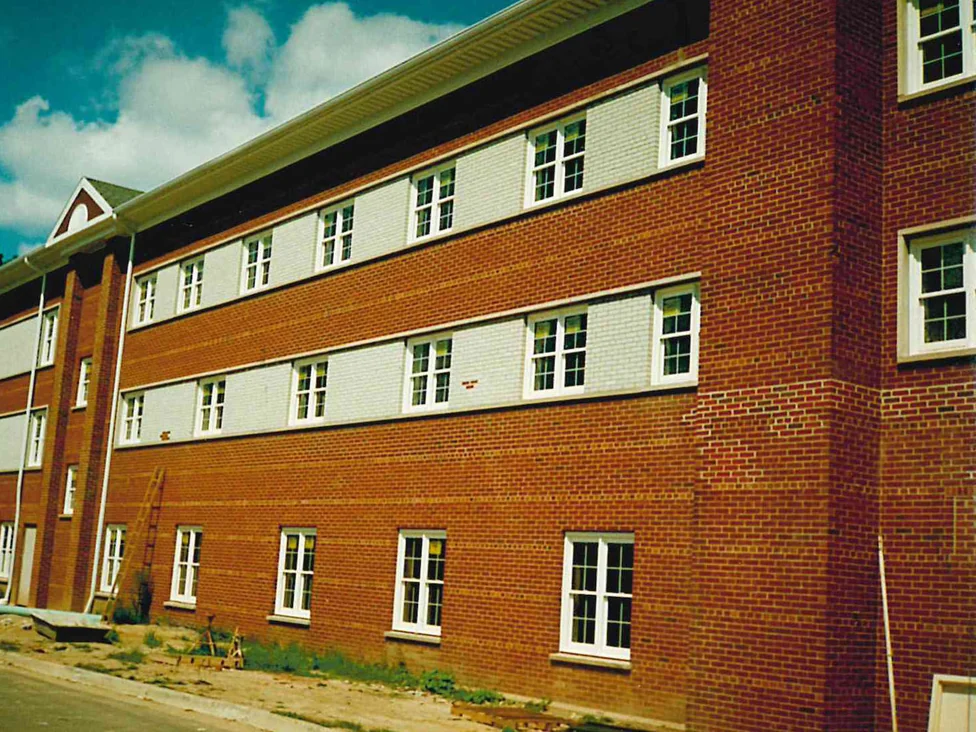A three-story building in red brick with two continuous bands of white brick carrying the upper windows, on bare ground before landscaping