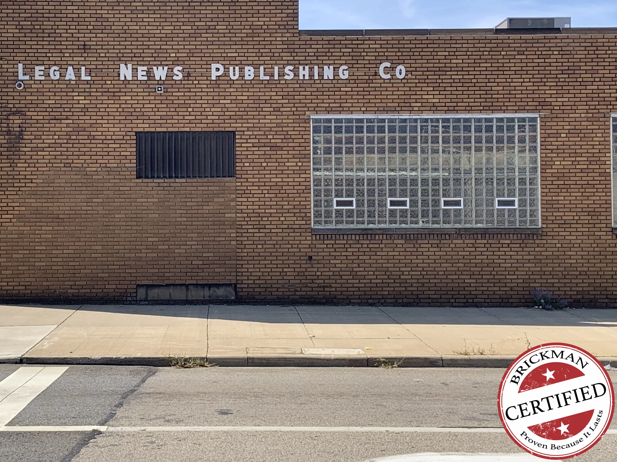 The office seen from across the street in 2019, its brick one red brown range from the sign band down to the sidewalk, with a glass block window bay to the right