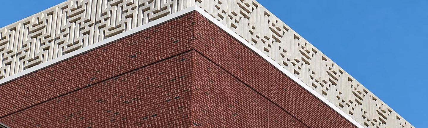 Upward view of a brick building corner topped with a white geometric concrete lattice cornice against a clear sky