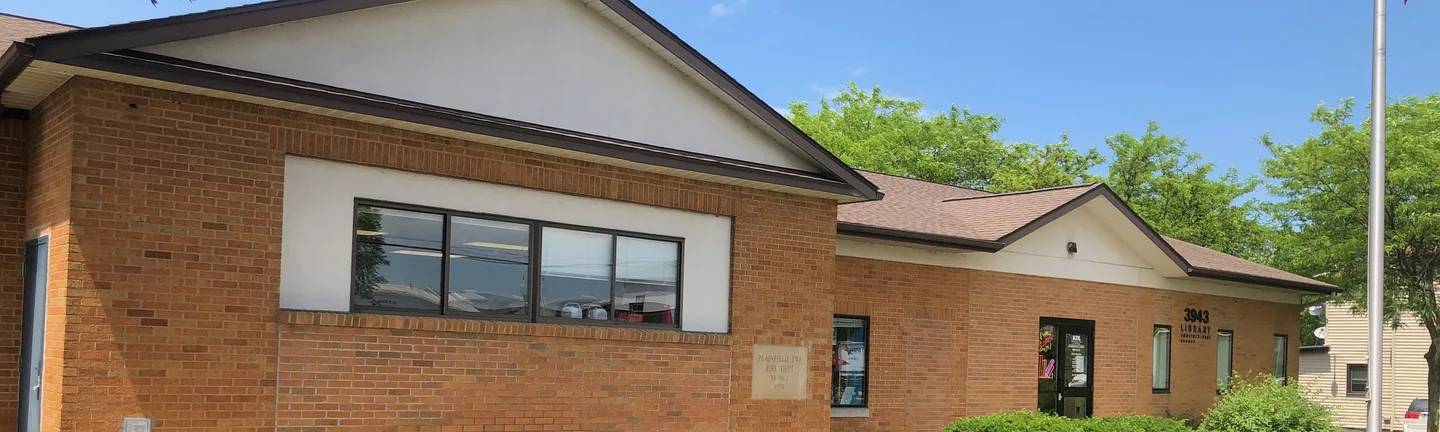 The Comstock Park branch library in 2019, a low run of tan brick under a shingled gable with a stone cornerstone beside the entrance