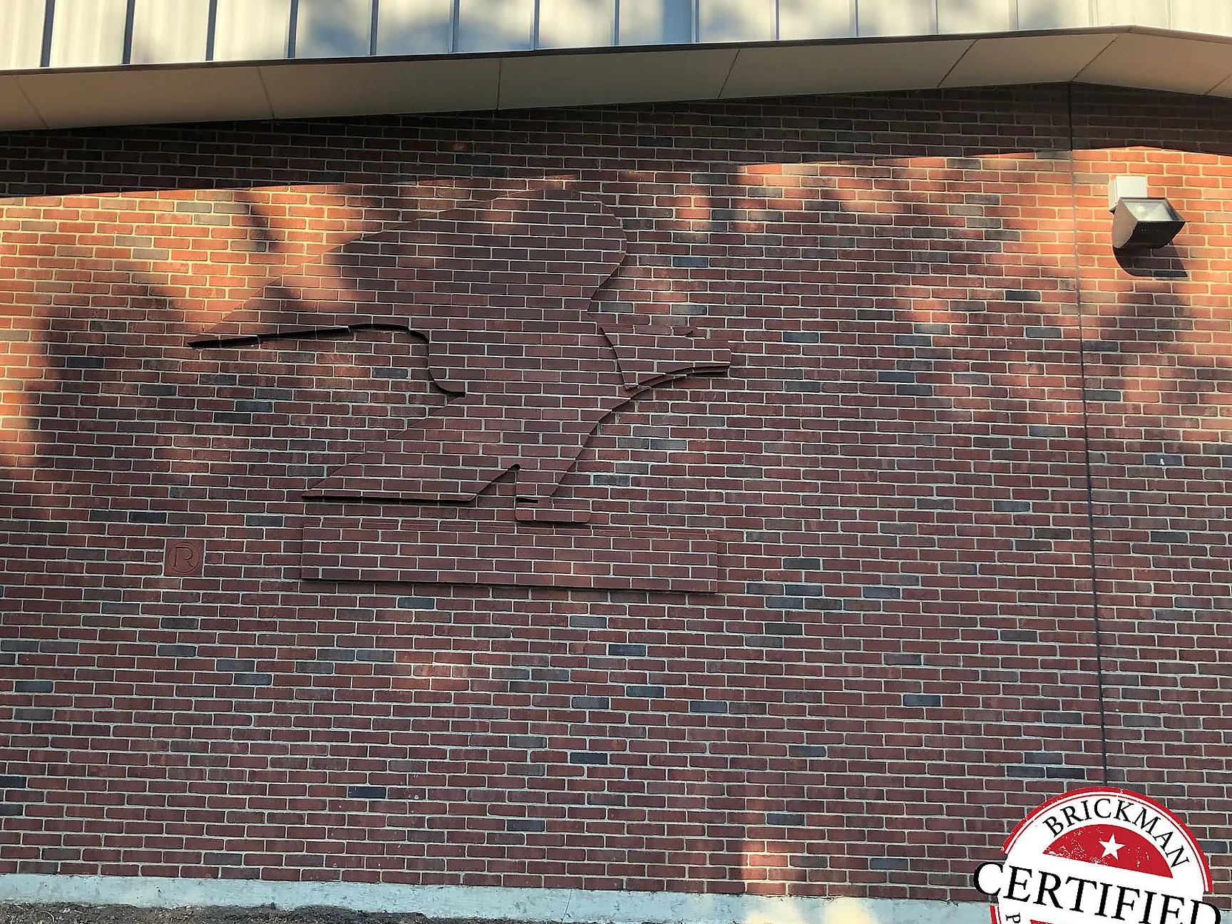The same eagle wall in 2019, sculpture and field brick reading as one dark red range under dappled tree shade