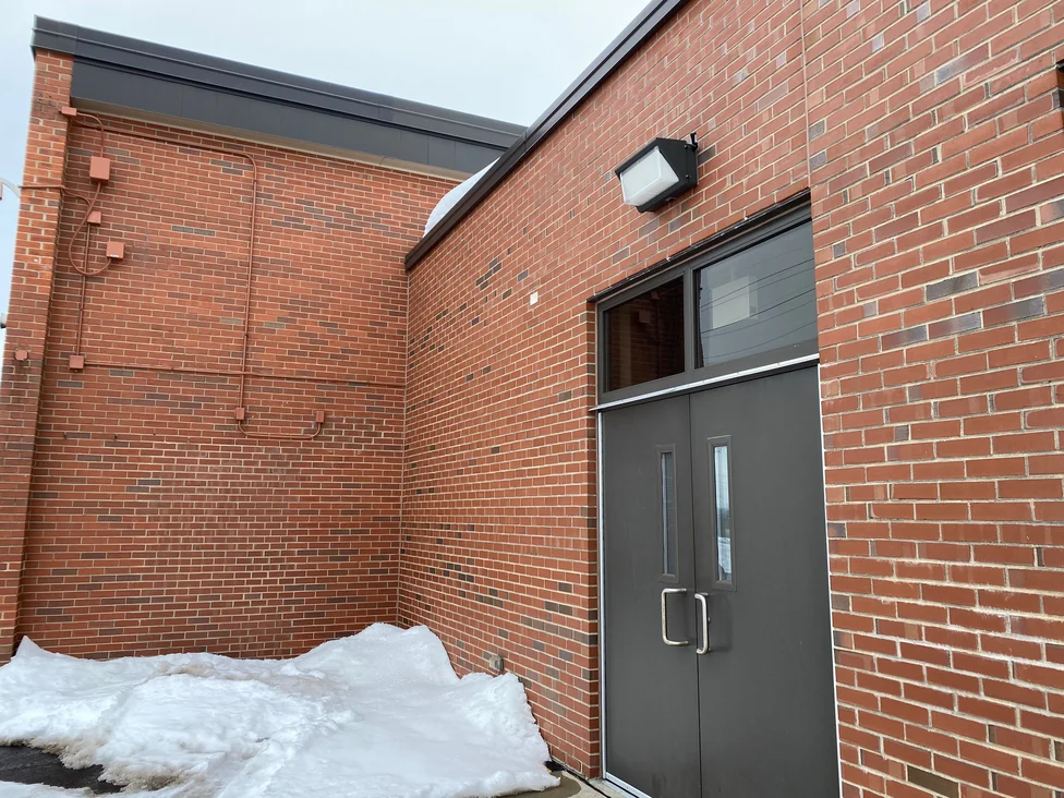 The same entrance in 2021 from close in and looking up, the lower wing meeting the taller wall at an inside corner, conduit painted to the brick on the left return and a snow drift against the doors