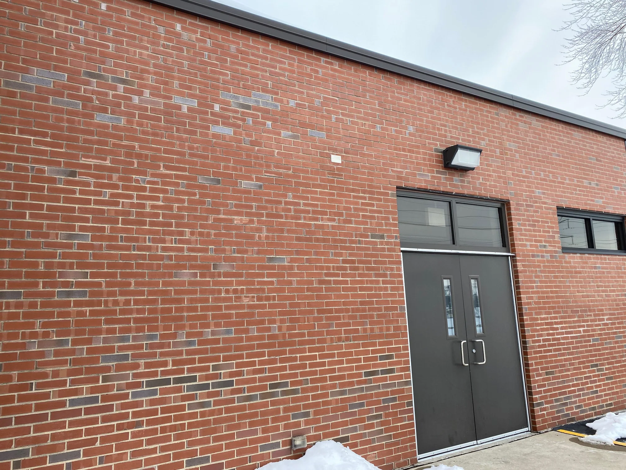 The entrance wall in 2021 seen from along the elevation, a long run of red brick with scattered grey headers carried past the dark double door to a ribbon window, snow and a yellow curb line at the foot of the wall