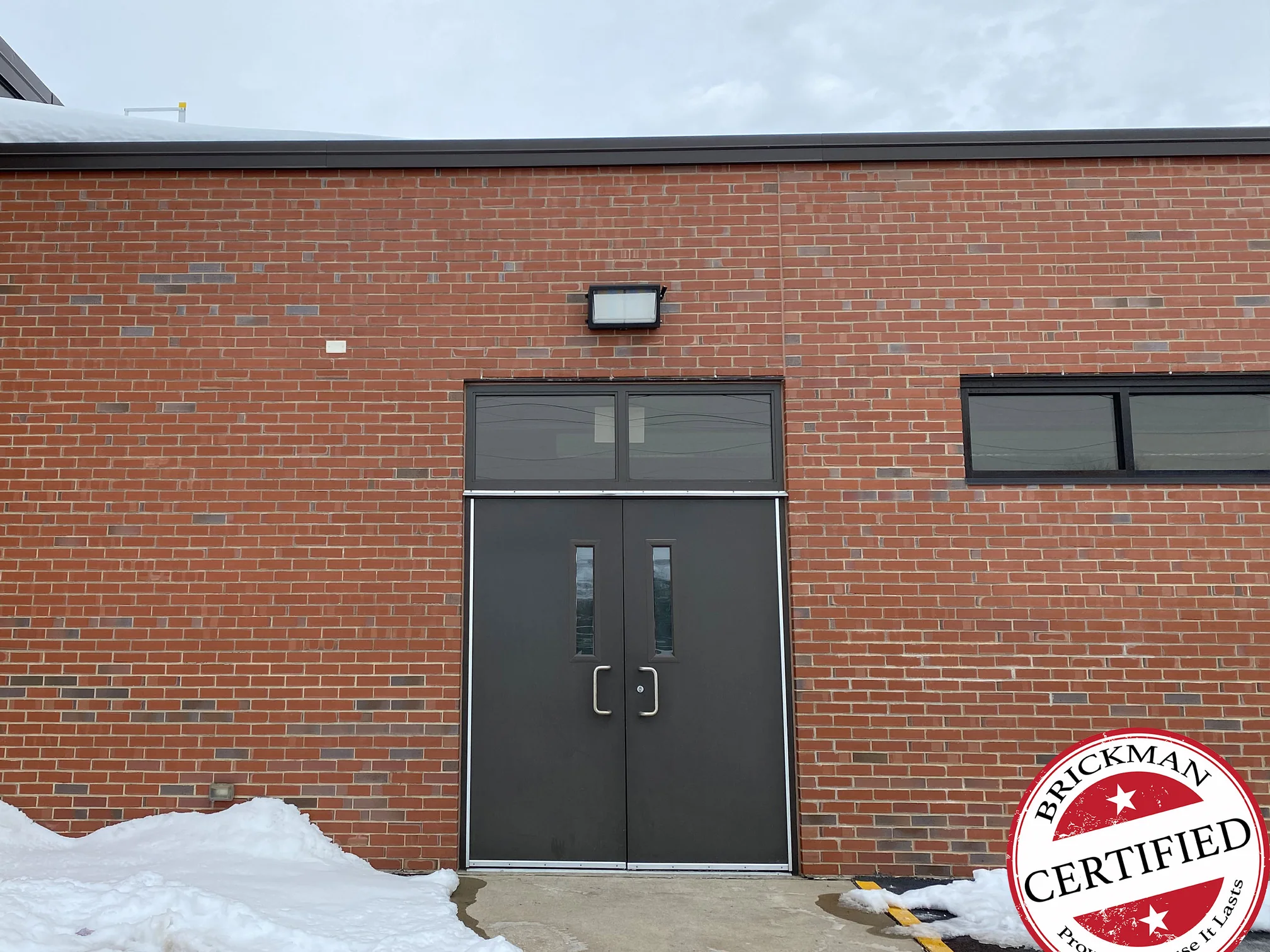 A school entrance in 2021, a dark double door with a transom set in red brick under a dark fascia, a ribbon window to the right, snow banked against the wall and the certification stamp in the corner