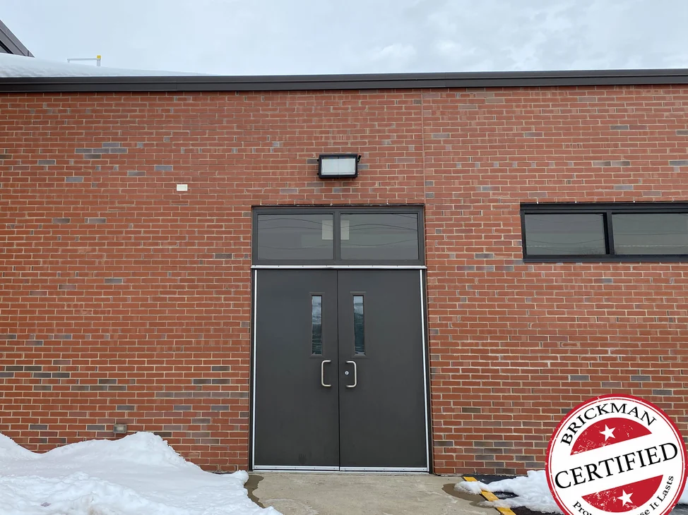 A school entrance in 2021, a dark double door with a transom set in red brick, snow banked against the wall and the certification stamp in the corner