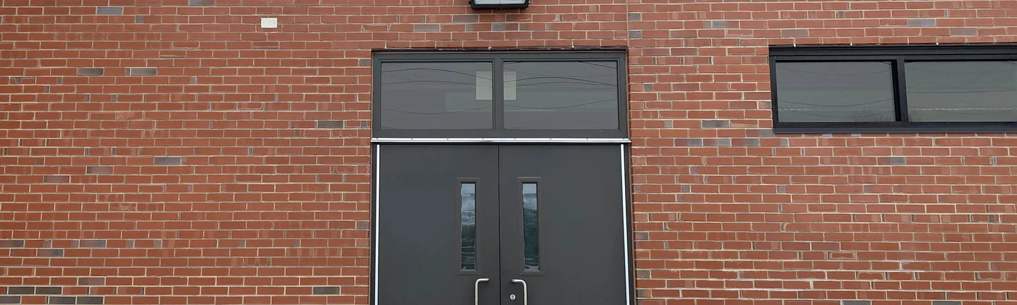 Red brick either side of a dark double door at Crest Hill School in 2021, a wall light above the transom and snow on the ground