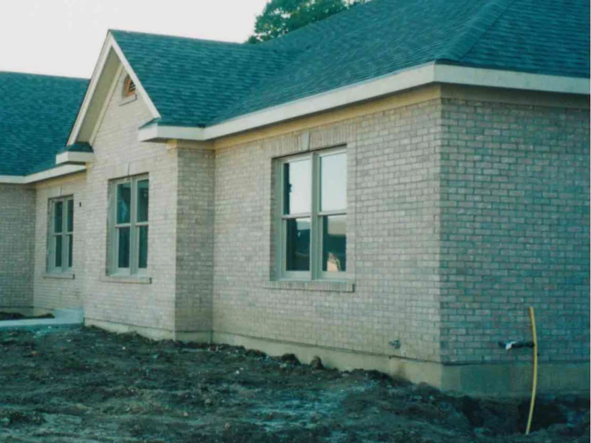 The corner of the house in October 1998 after the work, buff brick one even range around grey framed windows under a dark hip roof and a front gable, bare graded soil in front and a camera date of 10 10 98 in the corner