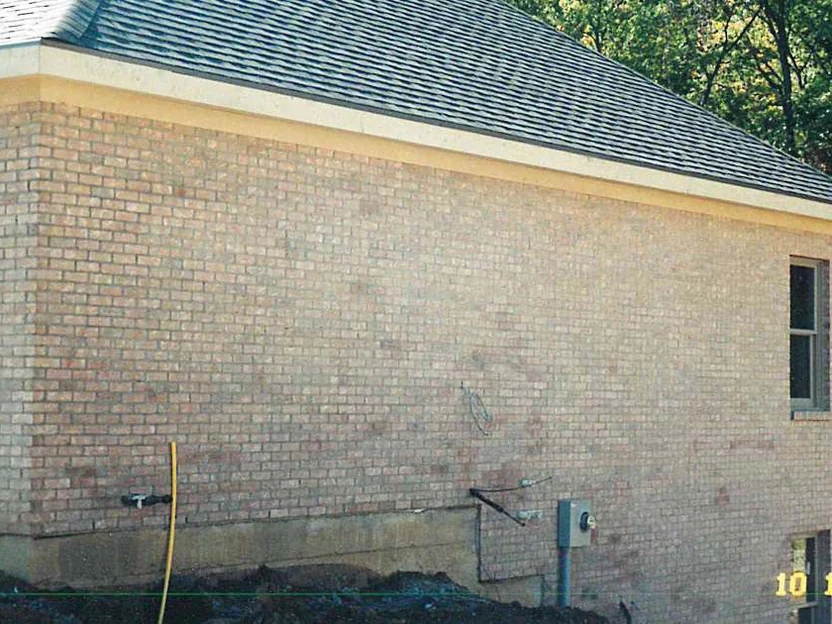 The side wall of the house in October 1998 before the work, buff brick with darker blotched units scattered across the lower courses, a hose and a meter box at the base