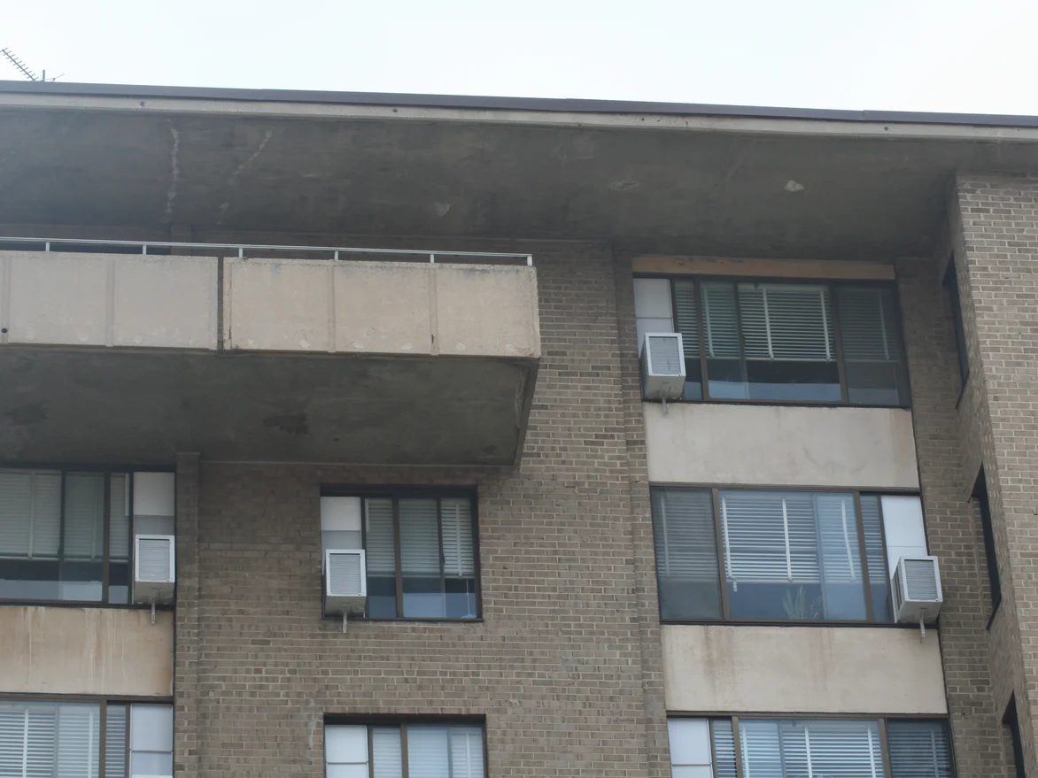 The top of the building in 2019, a deep concrete canopy carried over the upper floor windows with brick piers and cast panels beneath it