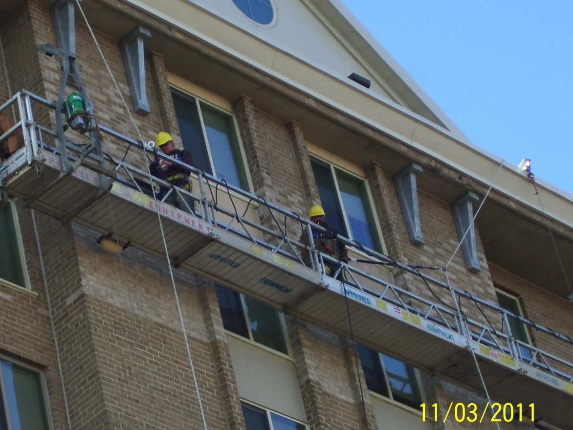 Two applicators in hard hats on a suspended work platform, working across the brick of the top floors below the gable