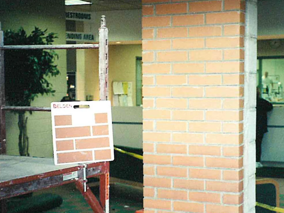 An interior brick column part way through the work, a Belden brick sample board on a stand beside it and scaffolding across the foreground