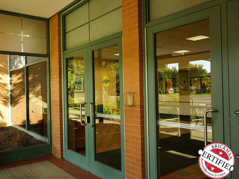 The glazed entrance in 2019, brick piers between the green framed doors in an even orange range, with the certification stamp in the corner