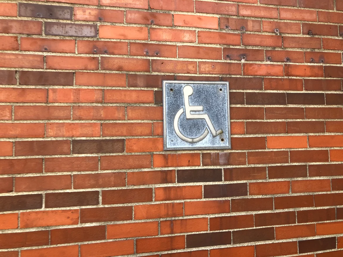 The ground level wall close up in 2021, red, orange and brown units laid in a mixed range with cream joints around a metal accessible plaque