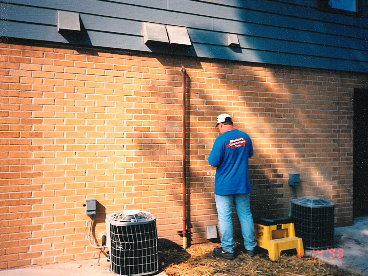 An applicator in a Masonry Cosmetics shirt working a filled-in section of the wall by hand, air conditioning units standing on the ground either side of him