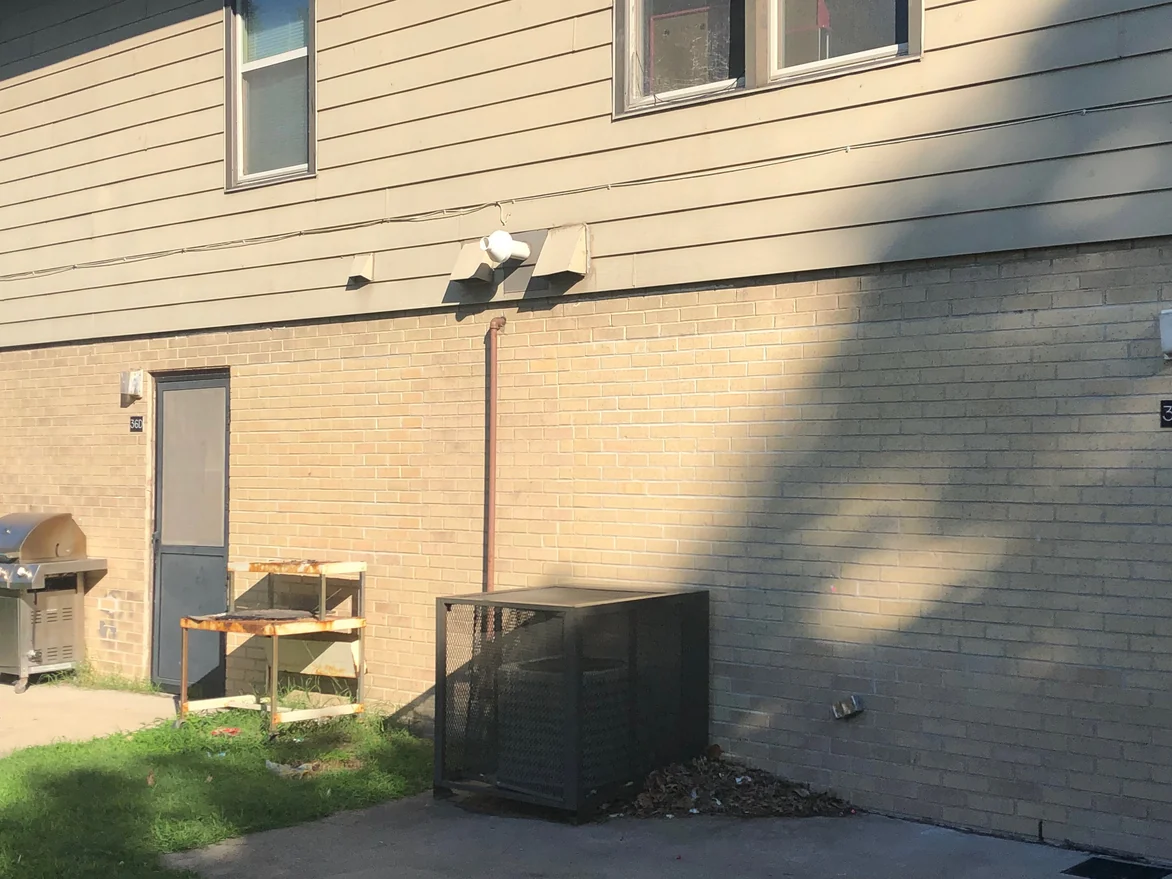 A rear elevation in 2019 with a screen door, a barbecue grill on the grass and an equipment cage on the concrete, the brick one buff range across the whole wall