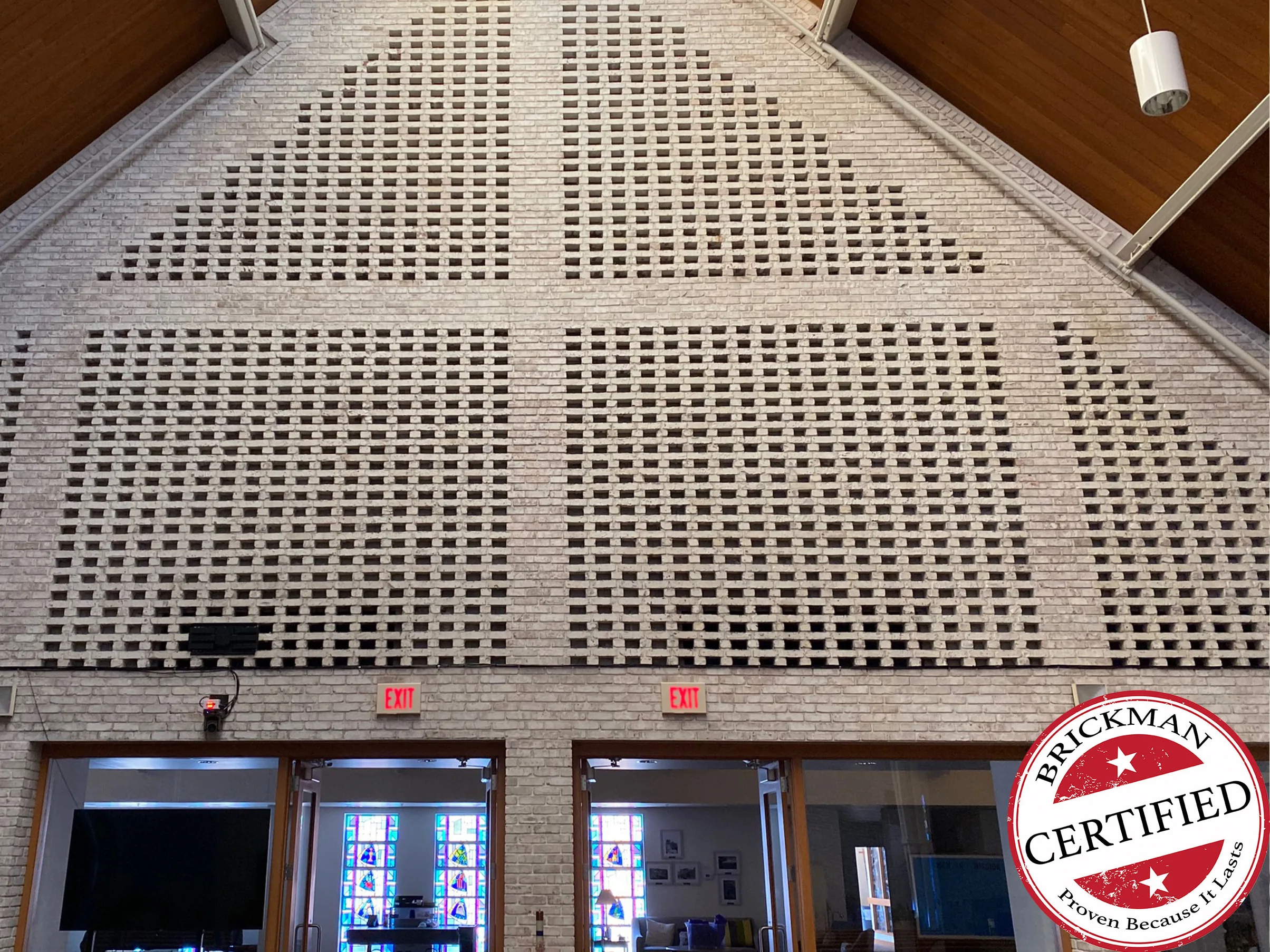 The gable end of the hall in 2021, one range of cream brick across the pierced screen wall above two pairs of doors, pendant lights hanging from the wood ceiling, with the certification stamp in the corner