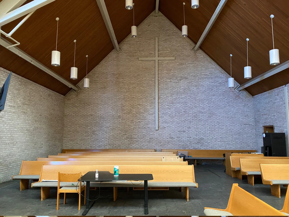 The hall in 2021 looking down the pews to the end wall, pale cream brick carrying a plain wood cross up to the ridge, cylinder pendants hung from the wood ceiling