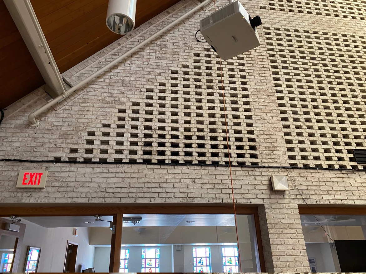 The pierced brick screen wall in 2021 seen from below, the open courses stepping down across it in the same cream range as the solid brick under them