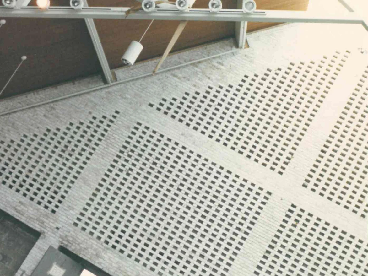 The pierced brick screen wall in 1989, shot from above through the roof trusses, pale brick with scattered darker units and a run of track lights rigged along the top