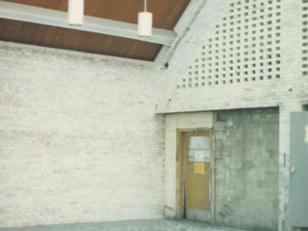 A side wall of pale brick in 1989 with darker units scattered through the courses, a single door in a block surround and the pierced screen wall rising to the right under a wood ceiling