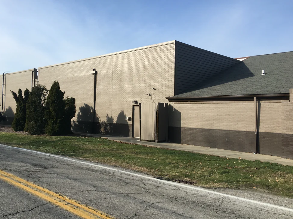 The long side wall of the Elyria restaurant in 2020, its brick under one grey tan finish with a darker band at the base and evergreen shrubs along the wall