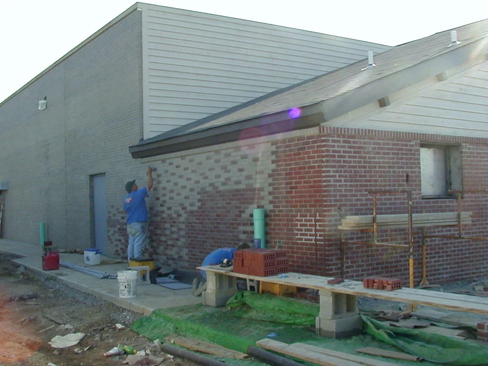 The light grey brick side wall of the restaurant in 2003 on the day staining began, an applicator brushing the wall beside a steel door while the front corner beyond him has already turned red