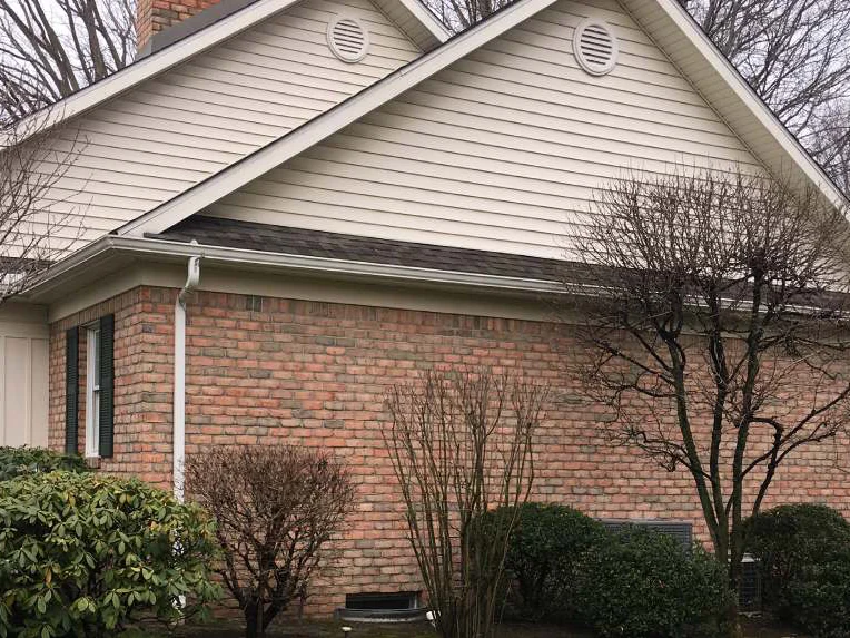 Two gables meeting in 2021, cream siding above and one range of red brick below, seen across a rhododendron