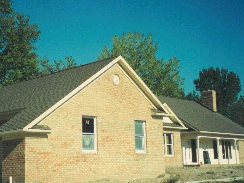 The gable end of the house in 1999, pale cream brick with scattered orange units under a round louvered vent, the ground still bare dirt from construction