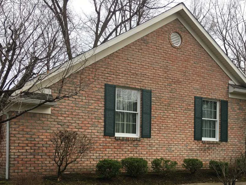 The gable end in 2021, the brick one range from the grade up to the round vent, two shuttered windows and a carriage lamp at the drive