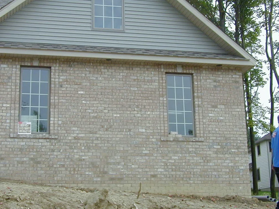 The side wall of a new two-story home in 2004, its light buff brick mottled with chalky white and grey units between two tall windows, bare earth graded in front