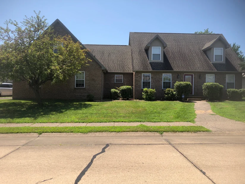 The house from across the street in 2019, the garage wing, the two dormers and the entry reading as one tan and brown range above clipped shrubs and a mature tree on the lawn.