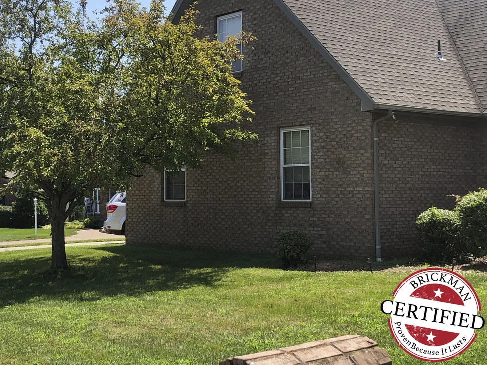 The gable end of the house in 2019 with a mature tree standing in front of it, the brick an even tan and brown blend from the lawn up to the gable window, and the Brickman certified stamp low in the frame